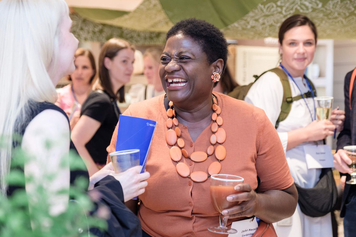 Attendee in orange top with statement necklace enjoys conversation at networking event with drink in hand.
