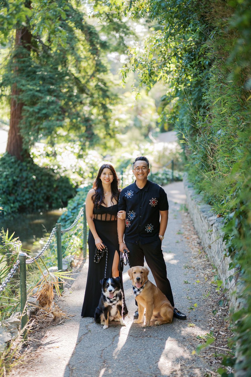 A couple with two dogs poses on a scenic garden path surrounded by lush greenery and trees.