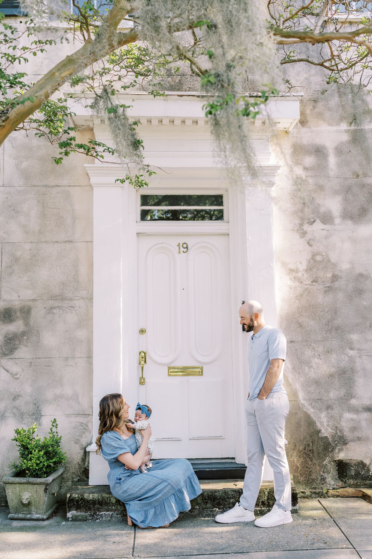 Two people in blue and white outfits sit and stand by a white doorway with Spanish moss and greenery in Charleston.