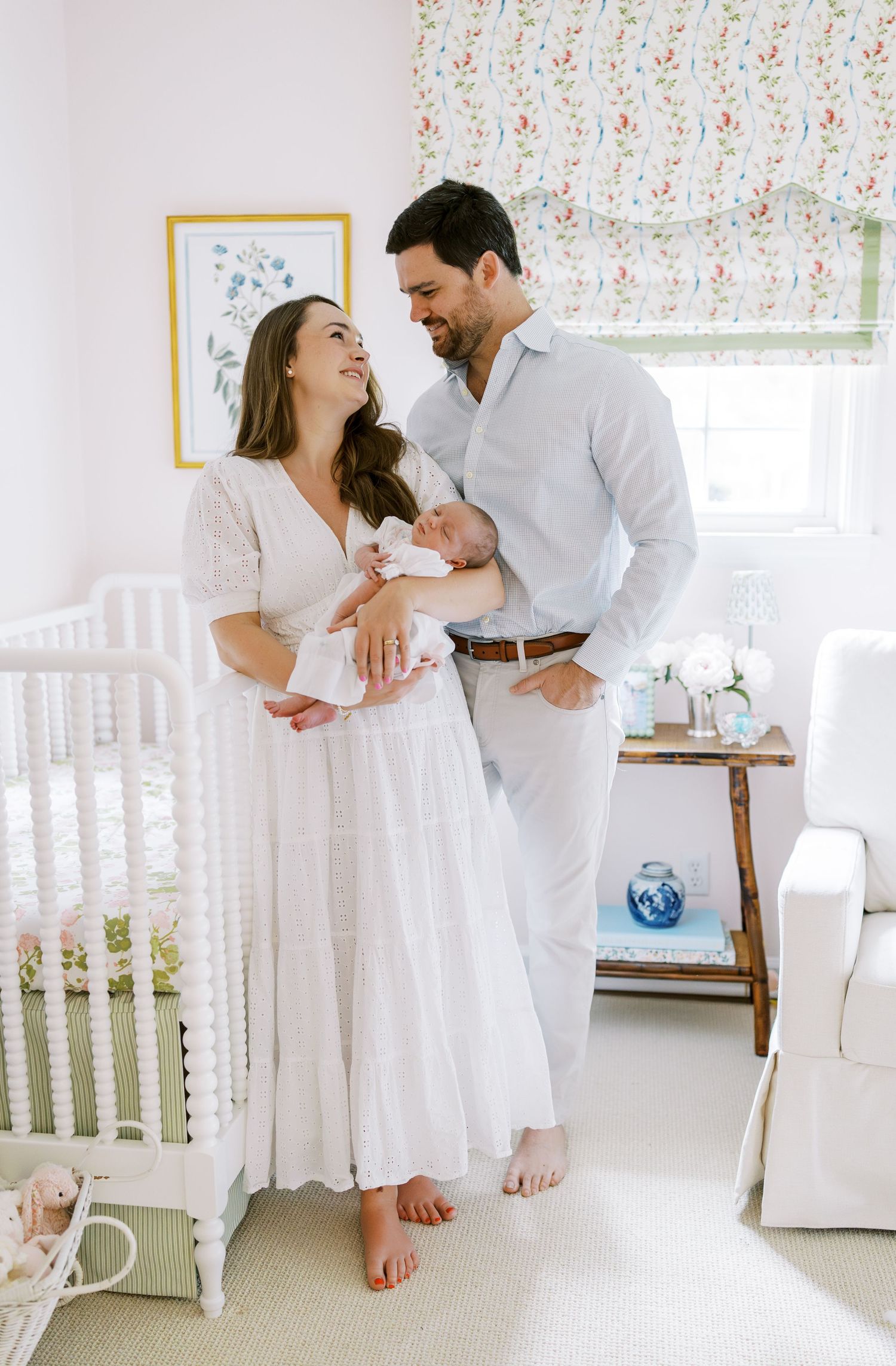 Couple wearing white stands together in a nursery with floral curtains while holding a newborn baby.