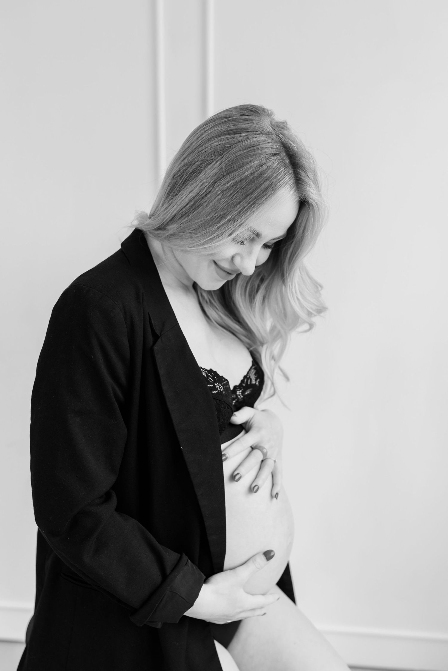 Black and white portrait of a person in a dark blazer posing intimately against a light wall.