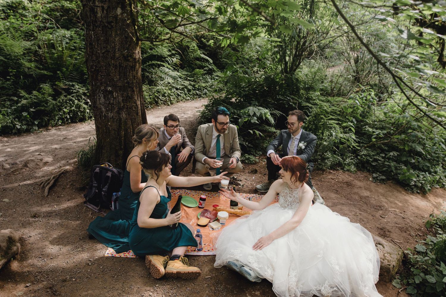 A wedding party enjoys a casual picnic together on a dirt path surrounded by lush forest greenery.