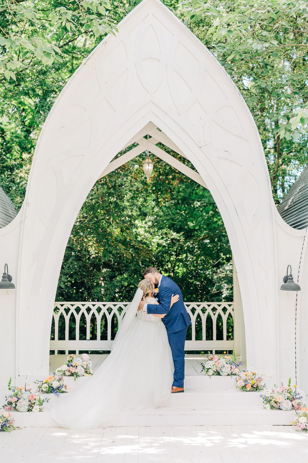 A romantic wedding scene under an ornate white gothic arch decorated with flowers in a garden setting.