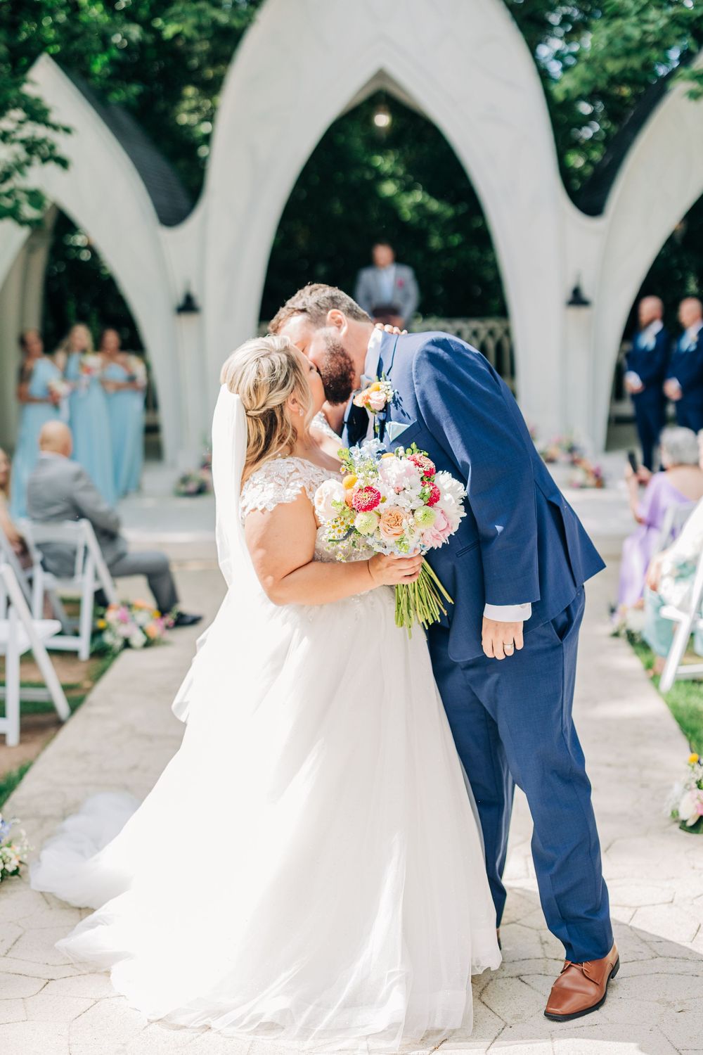 Newlyweds share their first kiss under ornate white arches while holding a pastel bouquet.