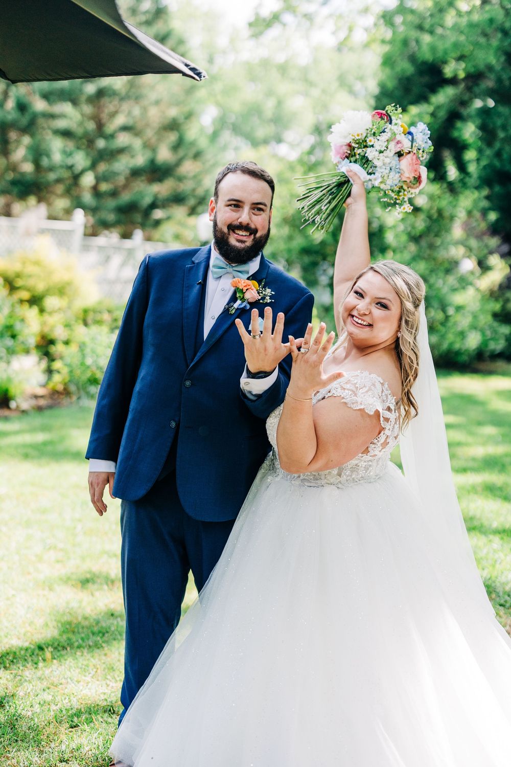 A wedding couple celebrating in a garden setting while showing off their rings and bouquet.