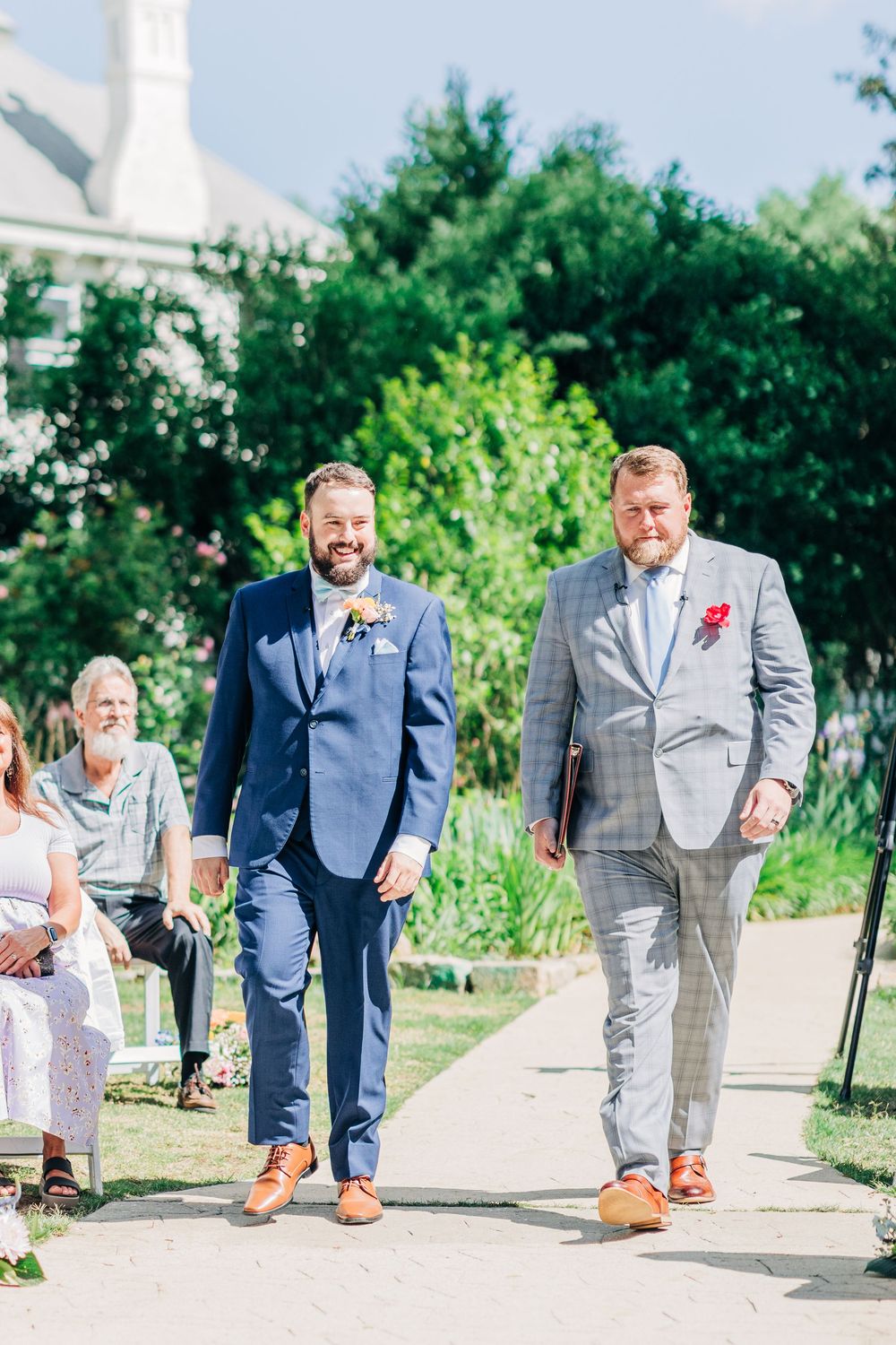 Wedding party laughs and walks together wearing blue and gray suits with brown shoes in an outdoor garden setting.