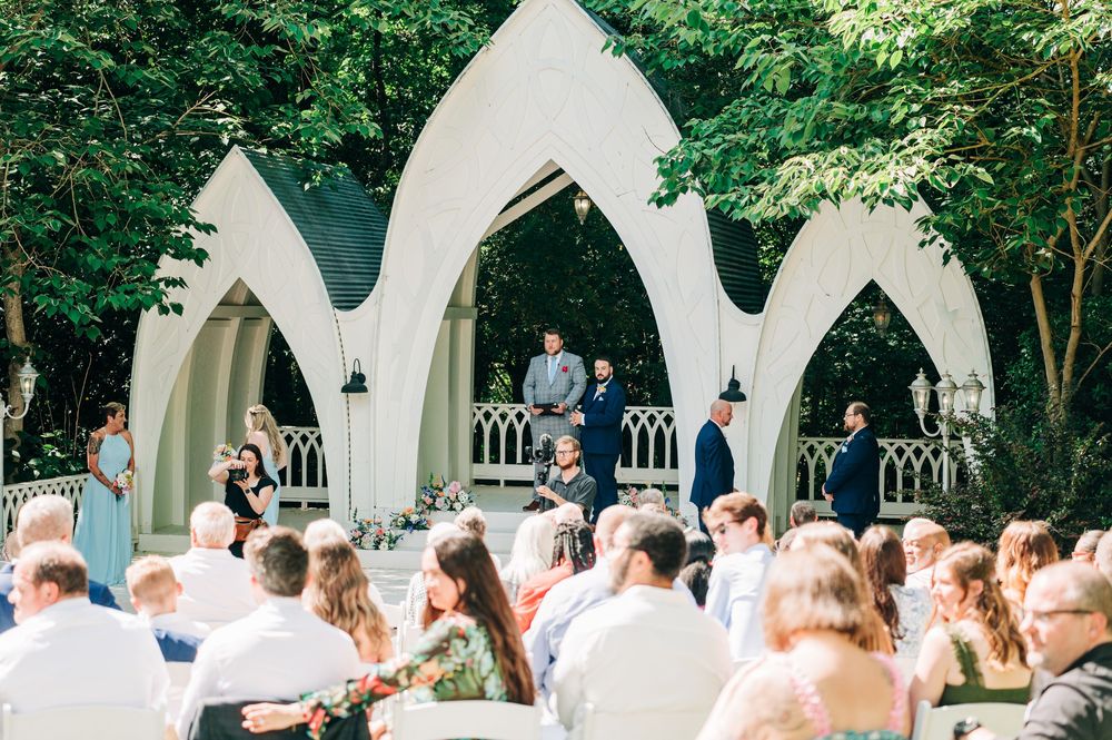 Outdoor wedding ceremony setup with white Gothic arches and rows of white chairs surrounded by lush green trees.