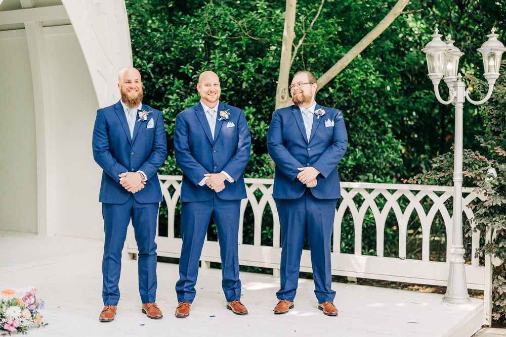 Groomsmen in navy blue suits and brown shoes sit on white railing with colorful garden background.