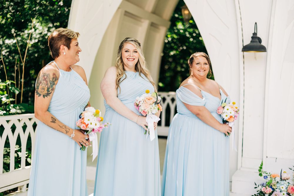 Three bridesmaids in matching light blue dresses hold colorful bouquets while standing on a white porch.