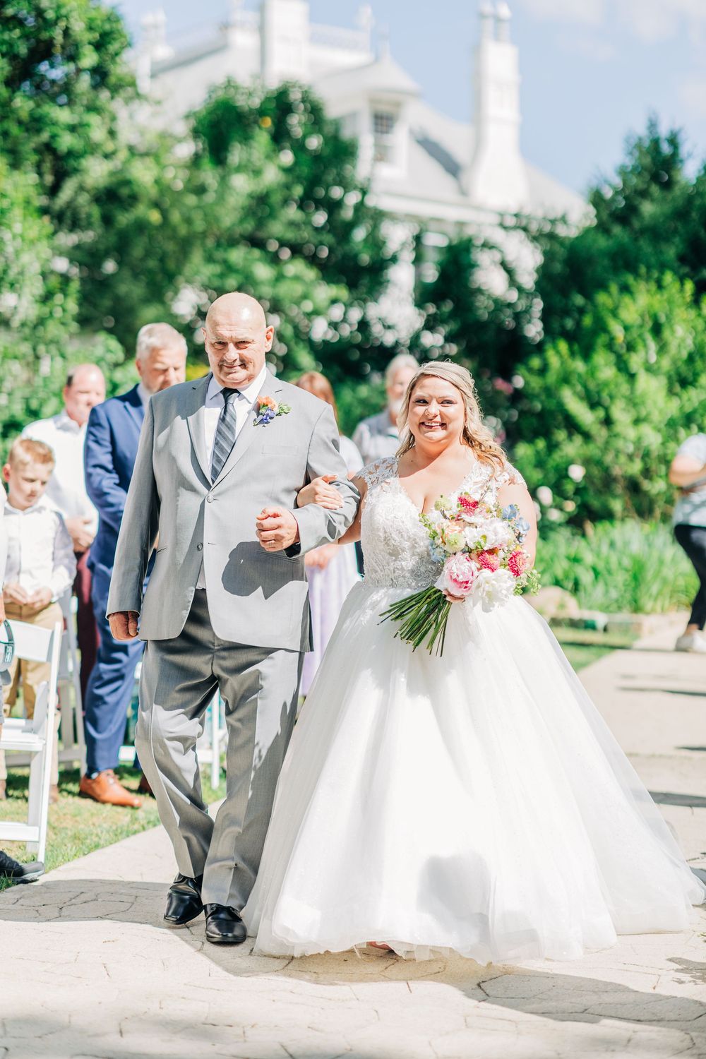 A wedding ceremony processional along a garden path with lush greenery and white architecture in the background.