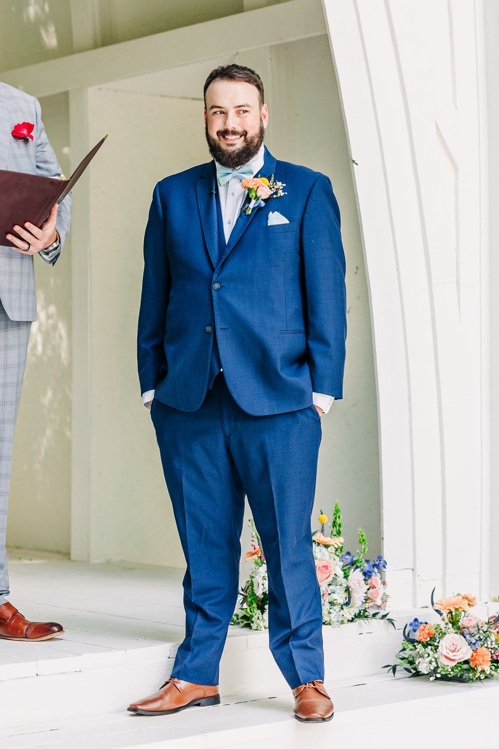 Men wearing navy blue suits stand in an elegant white columned space with floral decorations.