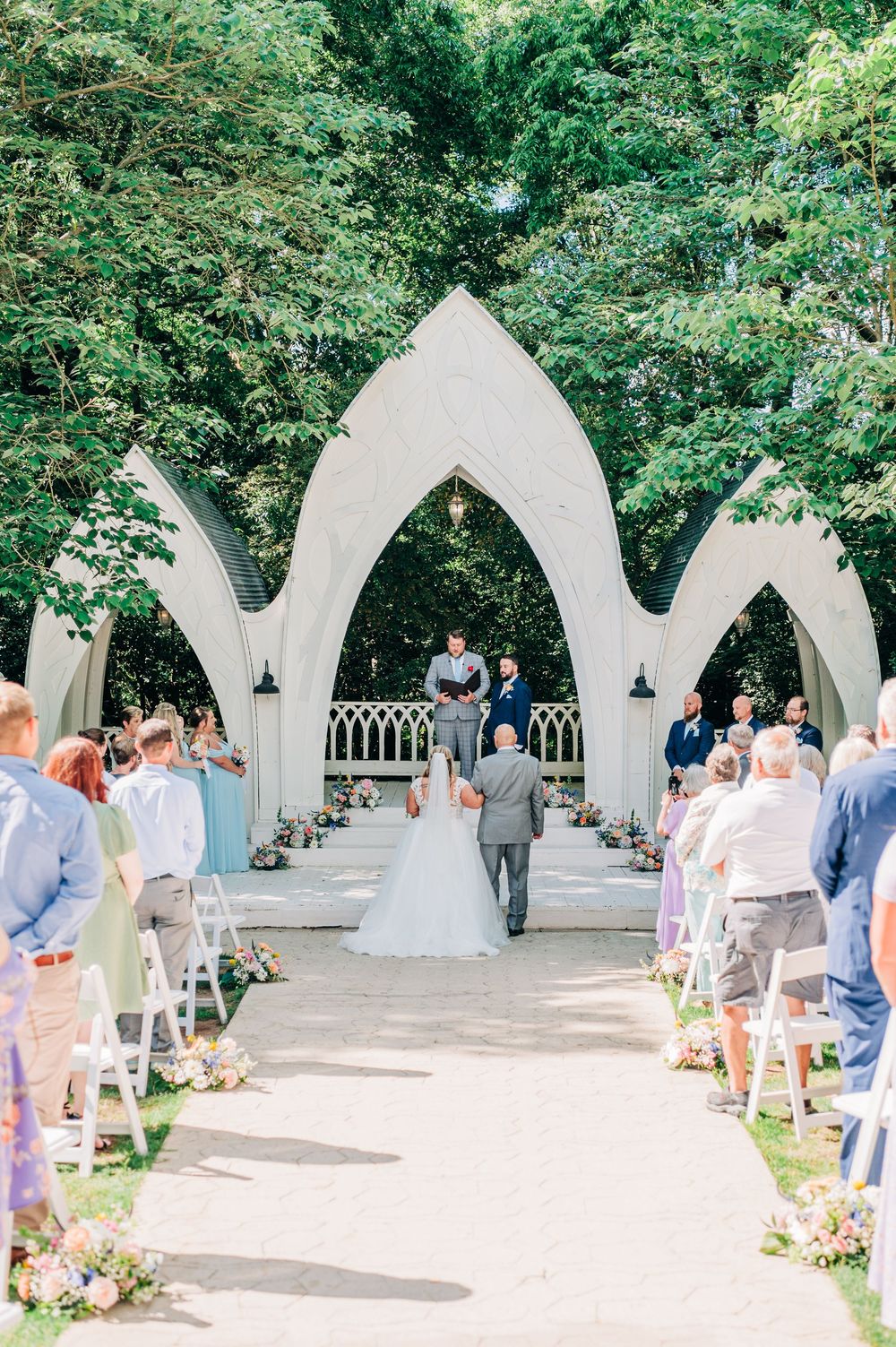 Elegant outdoor wedding ceremony at a garden chapel with white Gothic arches surrounded by lush greenery and guests.