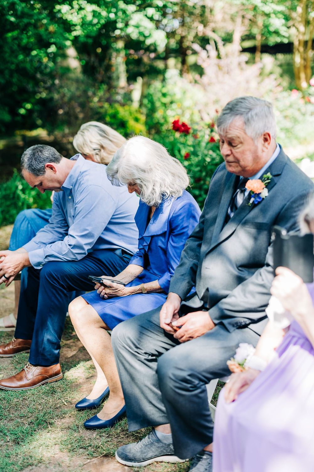 Wedding guests sit in a row outdoors checking their phones before the ceremony in a garden setting.