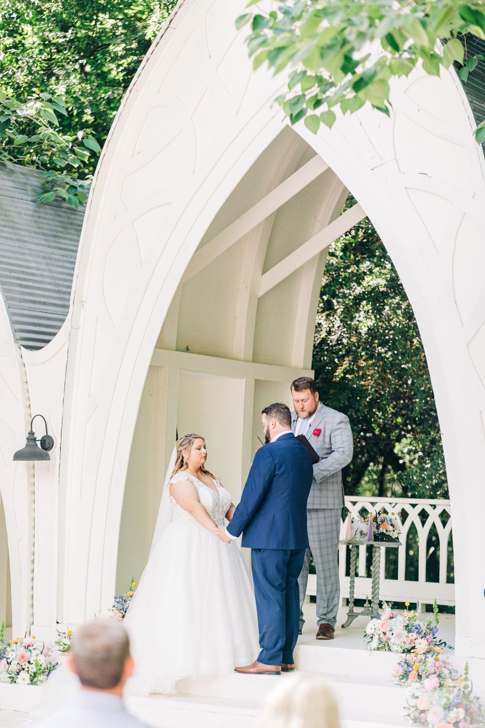 Outdoor wedding ceremony under a white arched gazebo surrounded by greenery and floral arrangements.