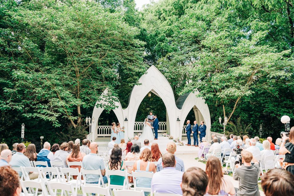 An outdoor wedding ceremony takes place under Gothic-style white arches surrounded by lush green trees and rows of white chairs.