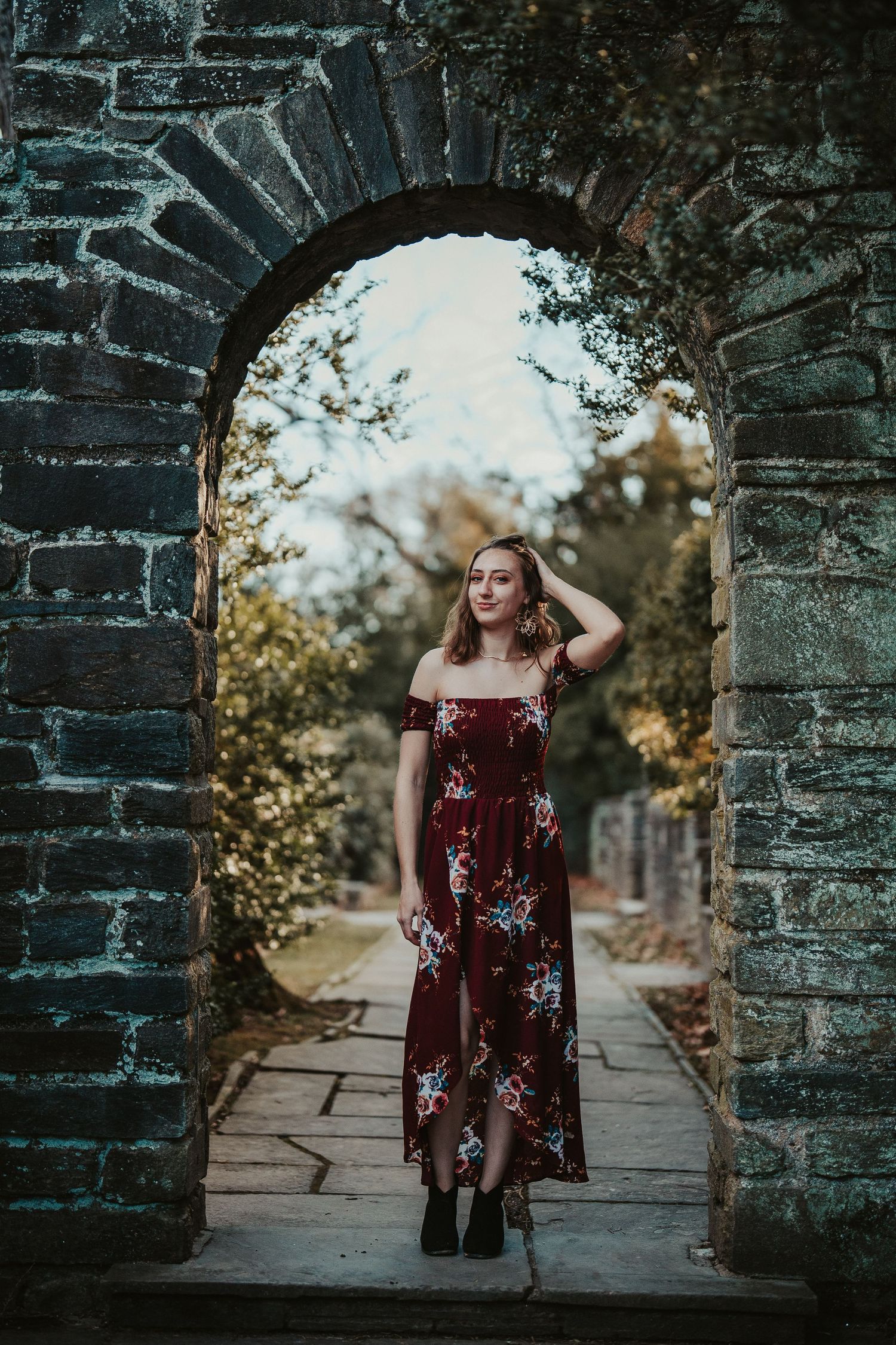 Figure standing in stone archway wearing burgundy floral off-shoulder maxi dress on stone path during golden hour.