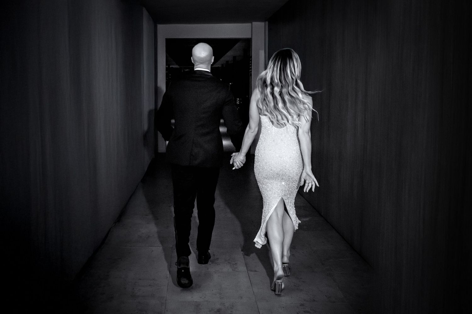 A couple walks down a dark hallway together, captured in black and white photography.