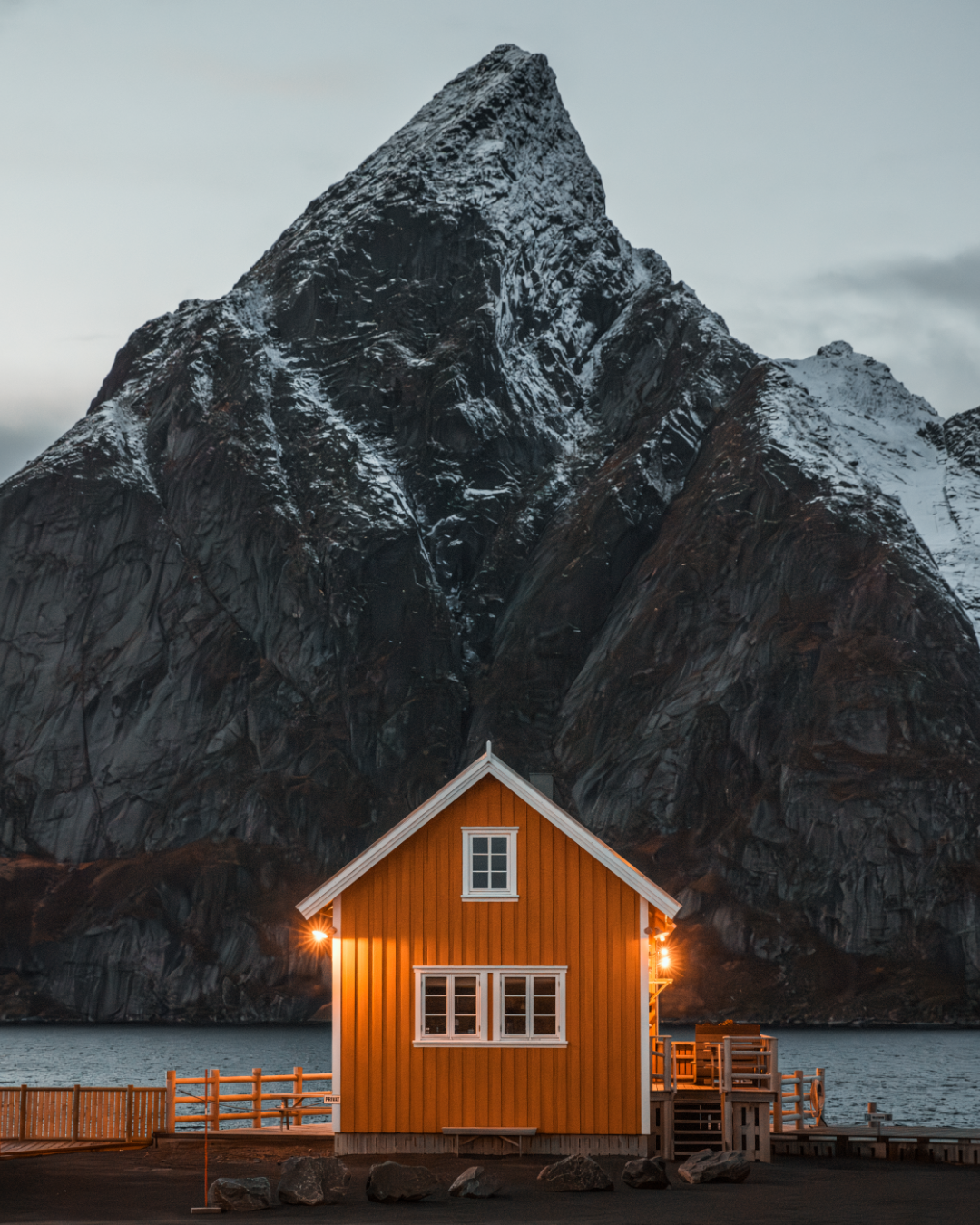 A cozy orange cabin glows against a dramatic mountain backdrop in Norway's Lofoten Islands at dusk.