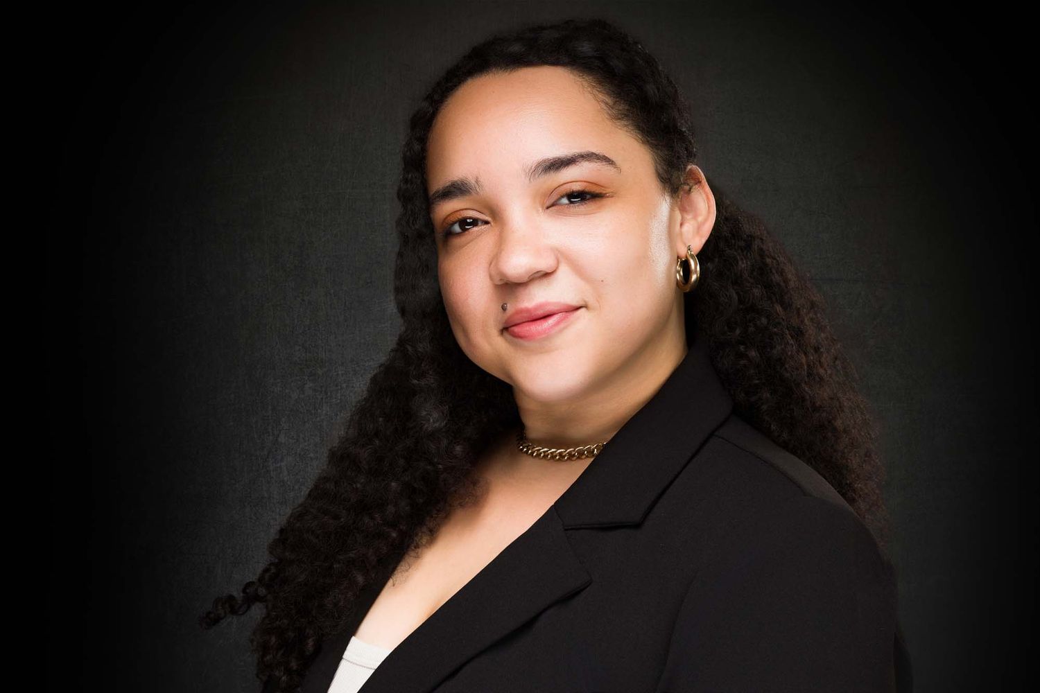 Professional portrait of a person with long curly dark hair wearing a black blazer against a dark background.