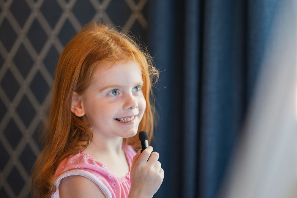 Young person with bright red hair and a pink top smiling while gesturing near a patterned wall.