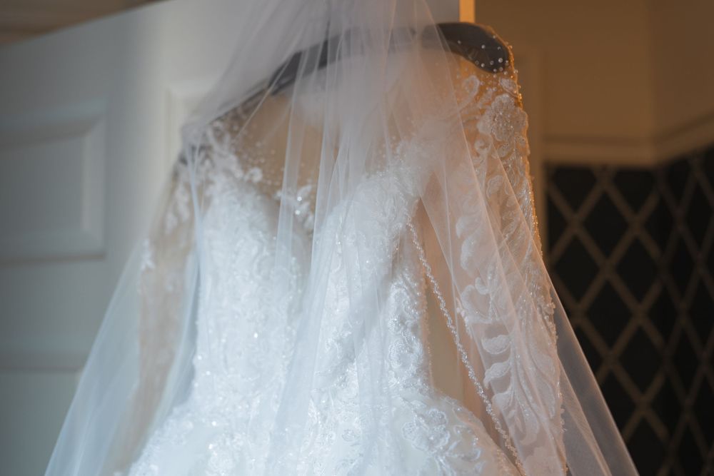 Close-up detail of a delicate lace wedding dress with veil hanging against a white door.