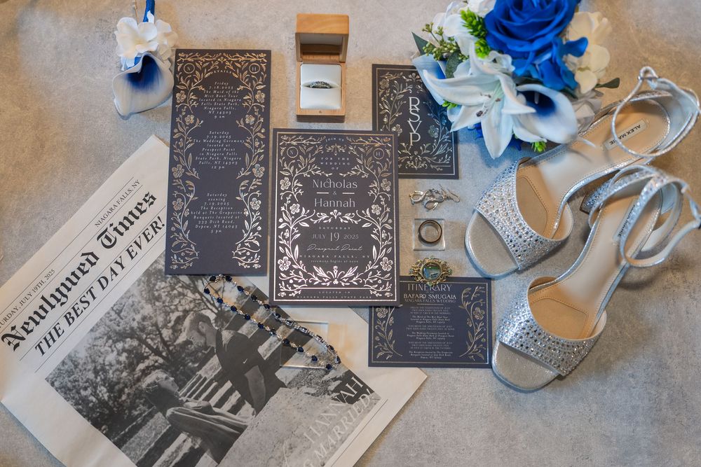 Wedding details flatlay with blue flowers, silver shoes, and vintage newspaper on gray background.