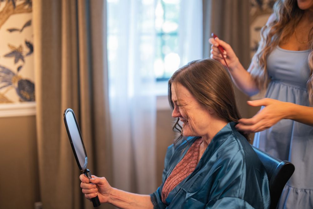 Woman getting her hair styled while looking at herself in a handheld mirror during wedding preparation.