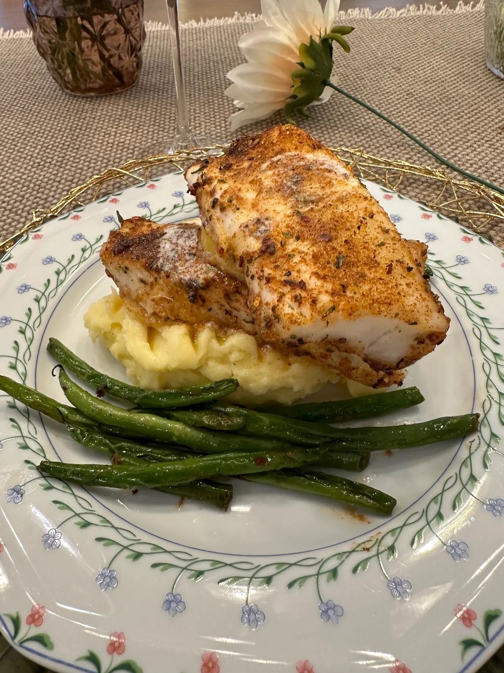 Seasoned baked chicken breast served with creamy mashed potatoes and sauteed green beans on a floral-patterned plate.