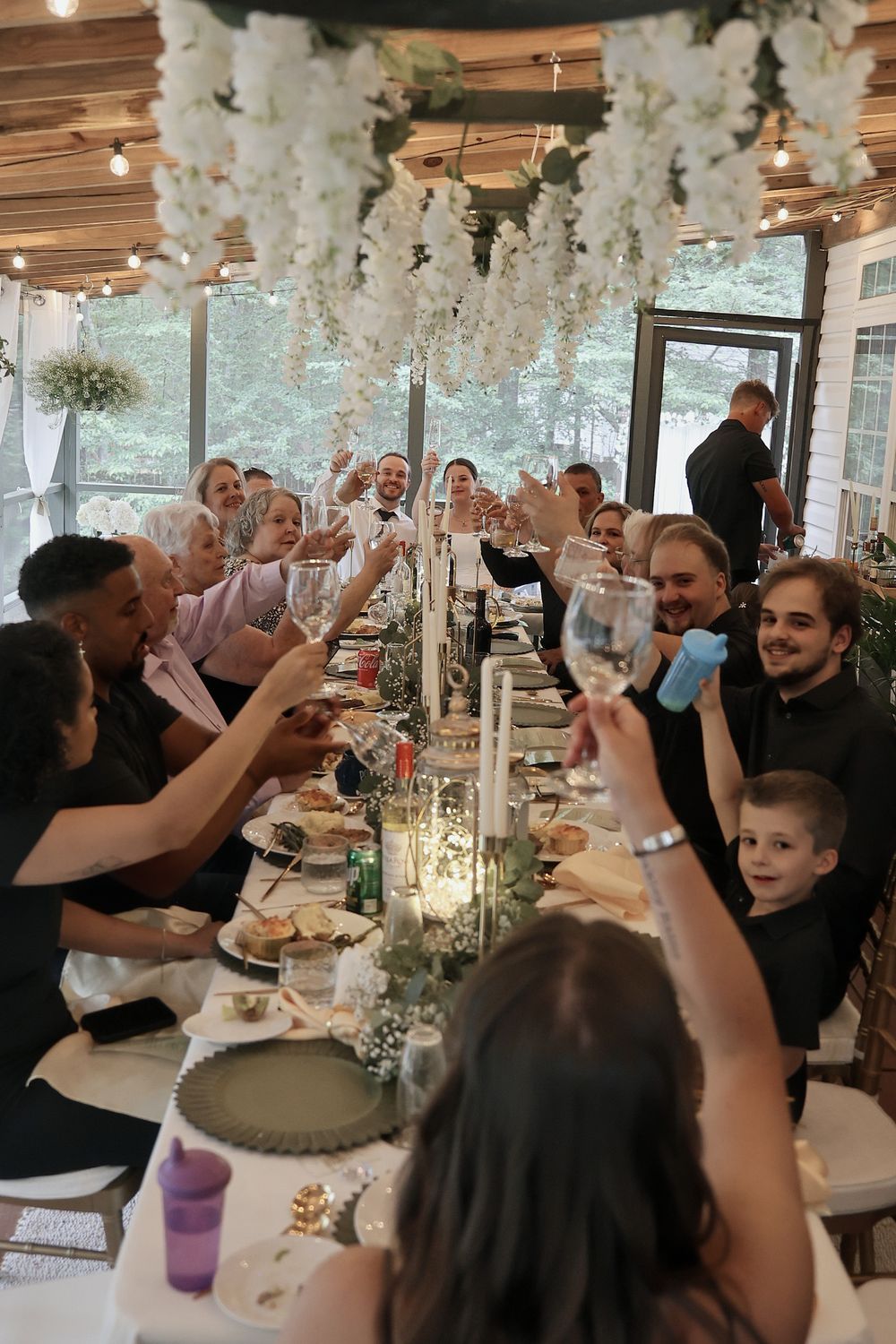 Wedding guests raise their glasses to toast during a reception under white floral decorations.