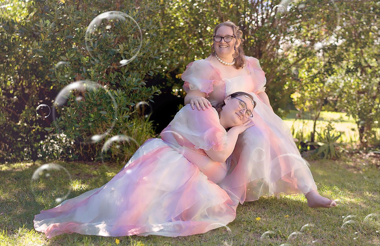 Two people in pink tulle dresses pose in a garden surrounded by floating soap bubbles in soft sunlight.