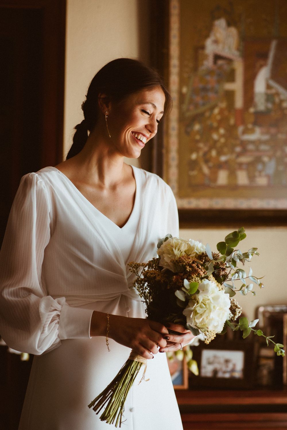 Bride in elegant white dress with flowing sleeves holding bouquet of white flowers while smiling.