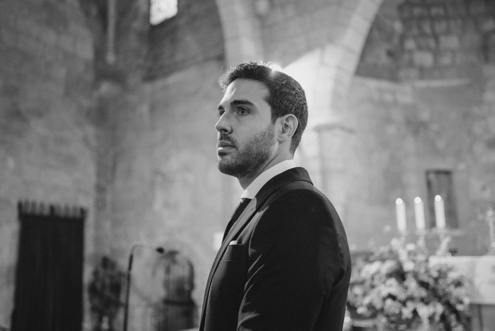 Black and white portrait of groom in dark suit standing in church with stone walls.