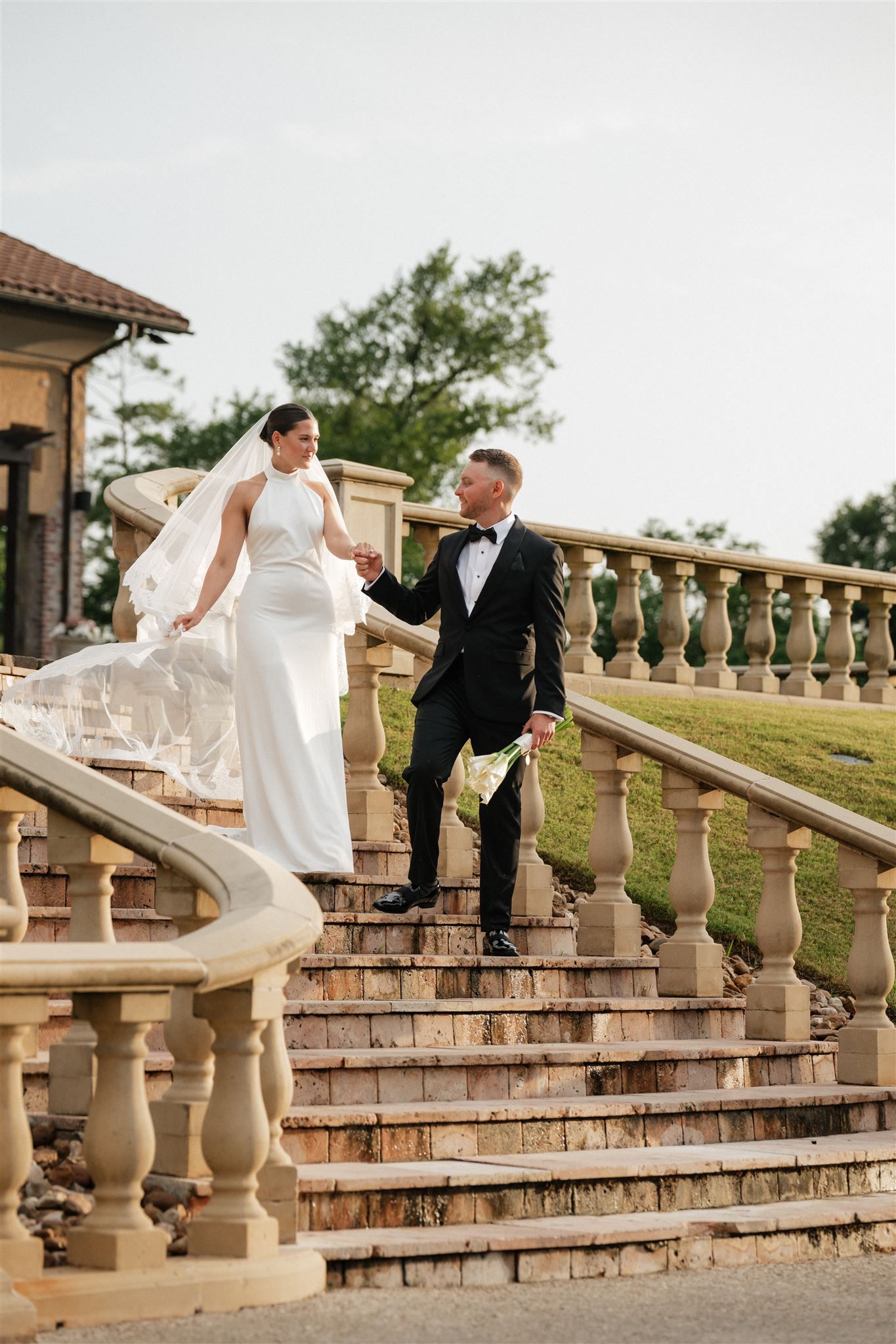 A couple in formal wedding attire descend an elegant stone staircase with ornate balusters at an outdoor venue.