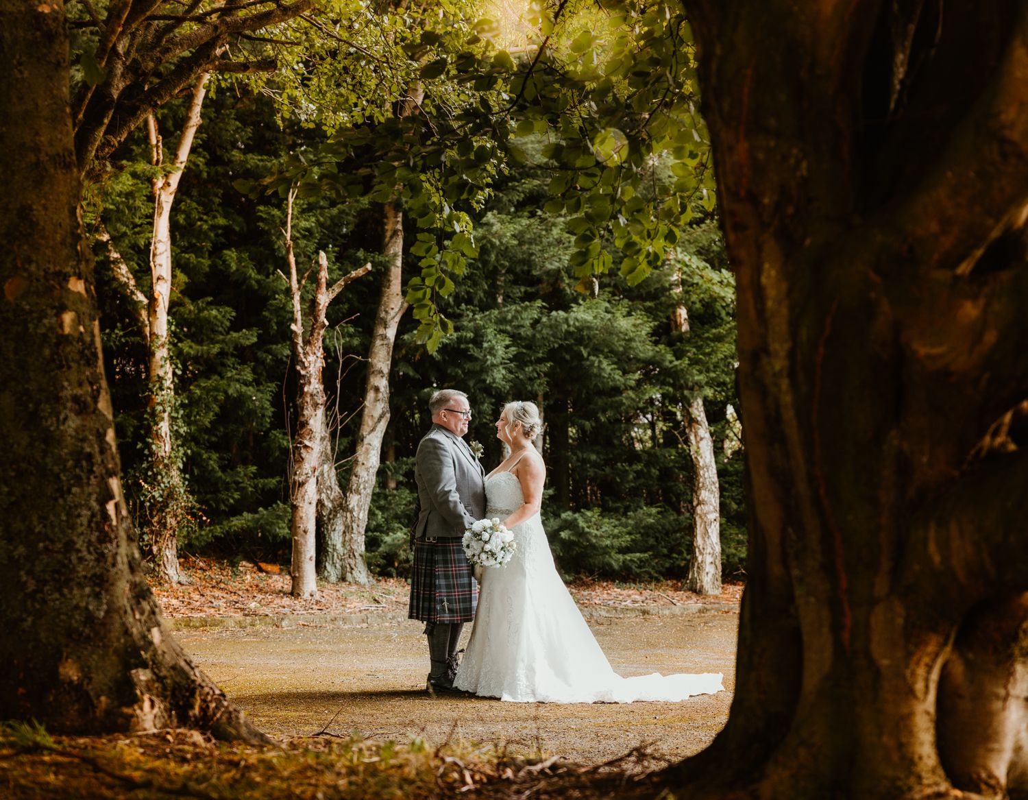 A romantic forest wedding scene framed by tall trees with sunlight filtering through the leaves on a wooded path.