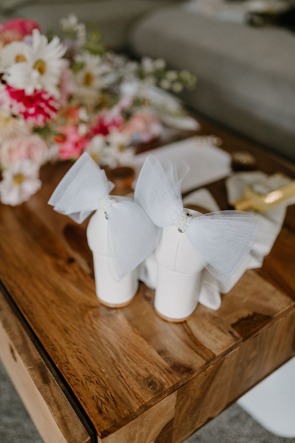 White bridal heels with delicate bows displayed on wooden surface next to pink and white floral arrangements.