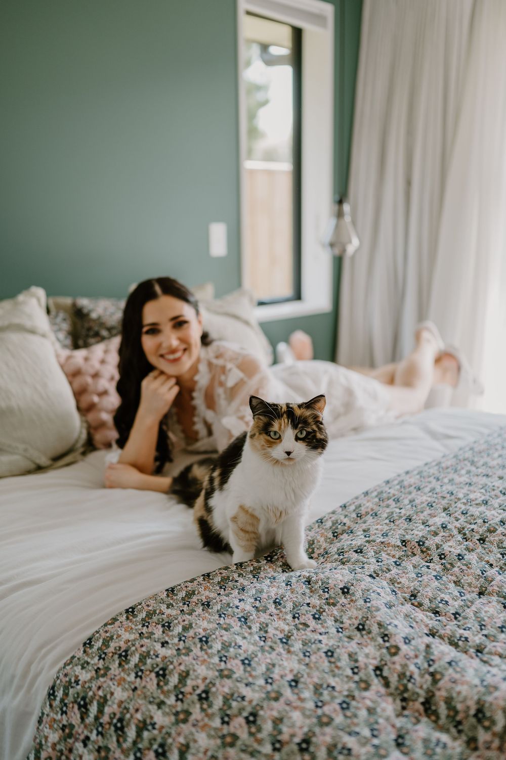 A black and white cat sits on a floral patterned bed with someone relaxing in a sage green bedroom.