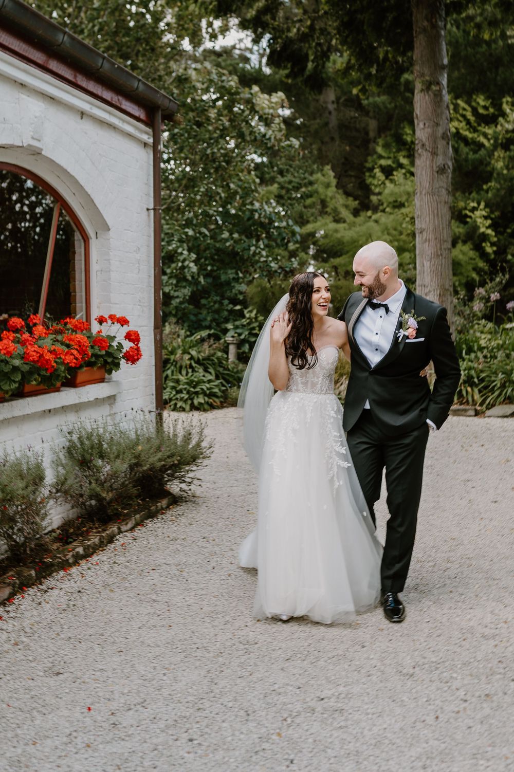 A couple walks along a garden path on their wedding day, with red flowers blooming in a window box nearby.