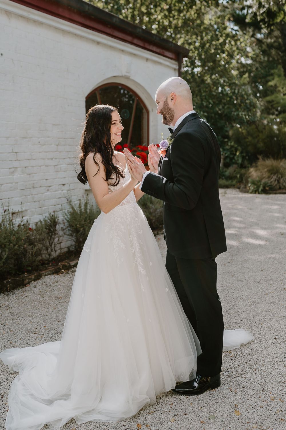 Romantic first look sequence between bride in white gown and groom in black suit on gravel path by white building.