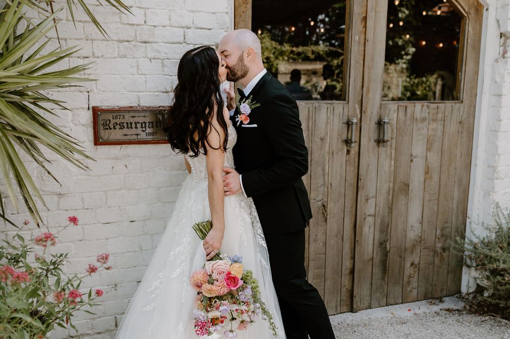 A romantic moment between newlyweds by a rustic door with palm fronds and pink flowers.
