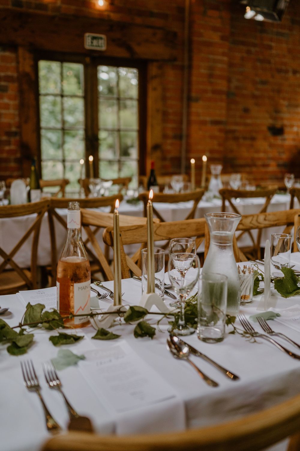 Wedding reception table with elegant place settings, wine glasses, candles and greenery decor in a rustic brick venue.