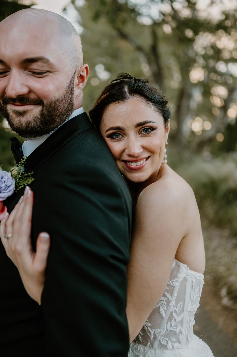 A wedding couple embraces outdoors in nature, with the bride in a strapless white dress and groom wearing a dark suit.