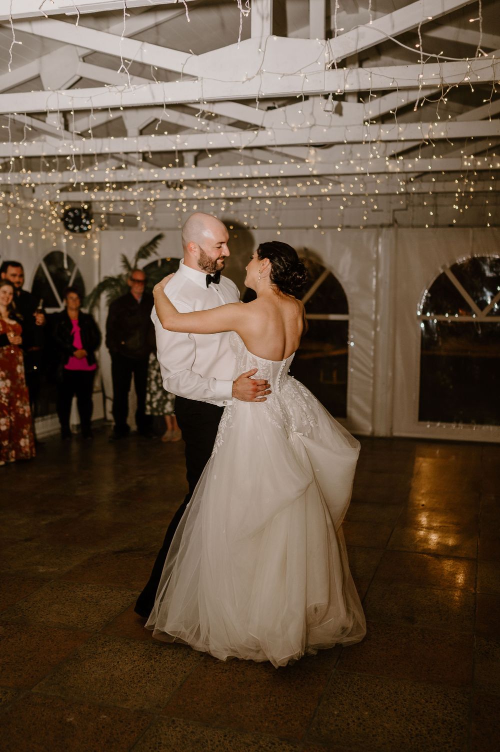 A couple shares their first dance at a wedding reception under romantic string lights and white ceiling drapes.