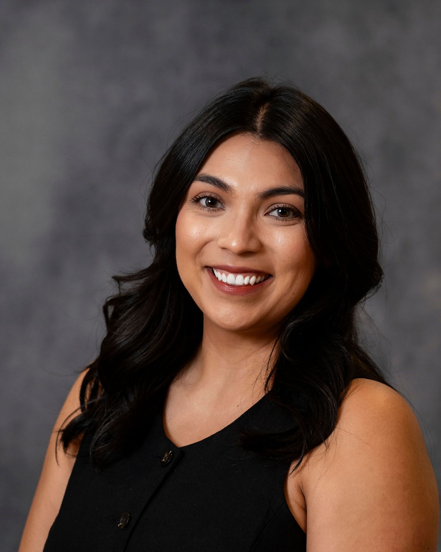 Professional headshot of a smiling woman with long dark hair wearing a sleeveless black top against a gray background.
