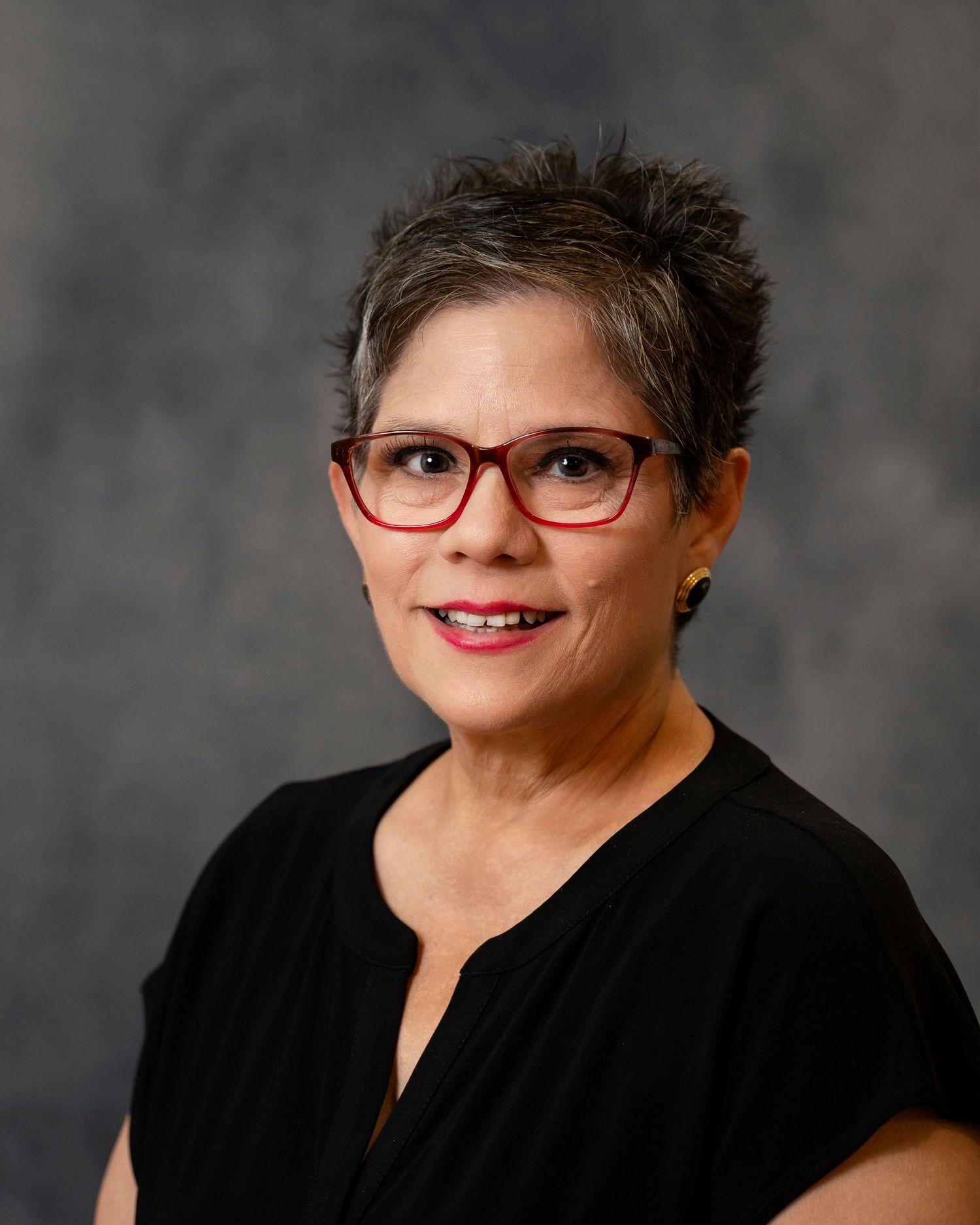 Professional headshot of a person wearing black top and red-framed glasses against gray background.