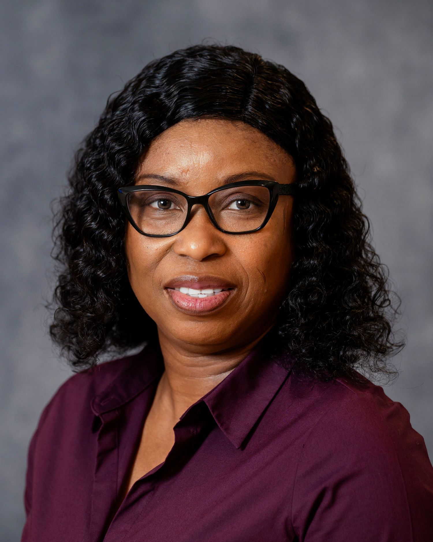 Professional studio portrait of a person in a burgundy blazer and glasses against a gray background.