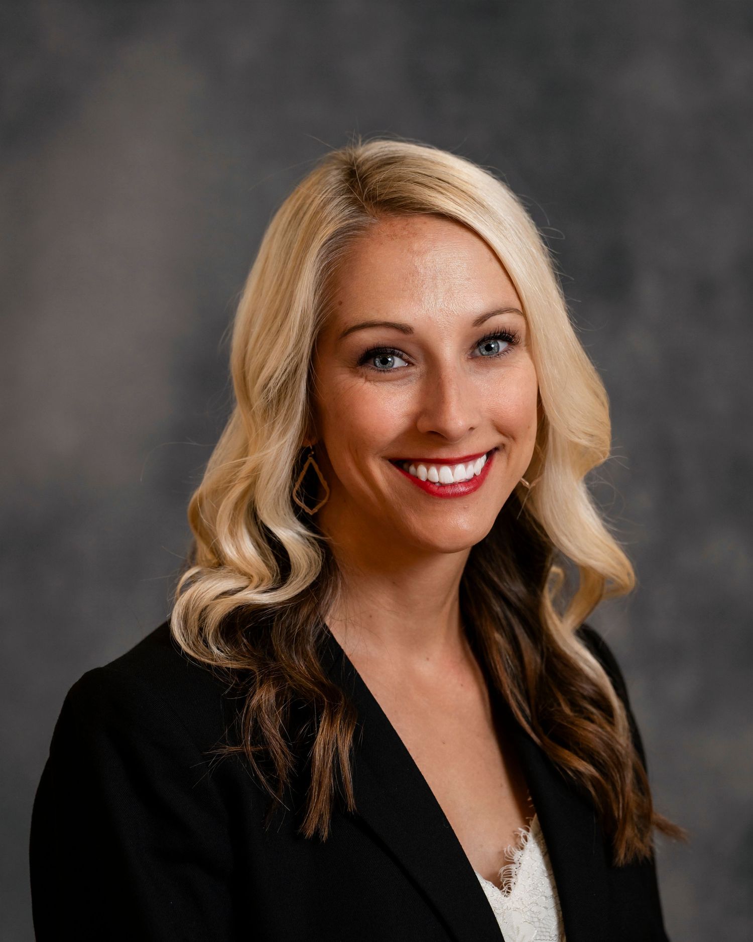 Professional portrait with blonde hair wearing dark blazer against gray backdrop.