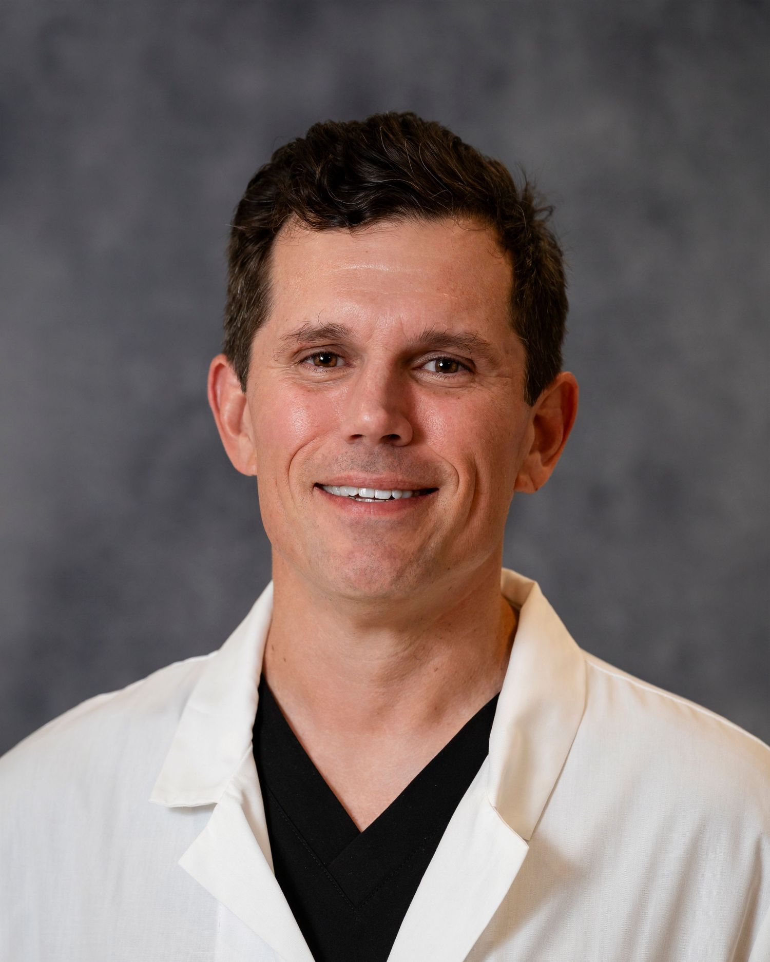 Healthcare professional in white coat and dark shirt smiling against gray background.