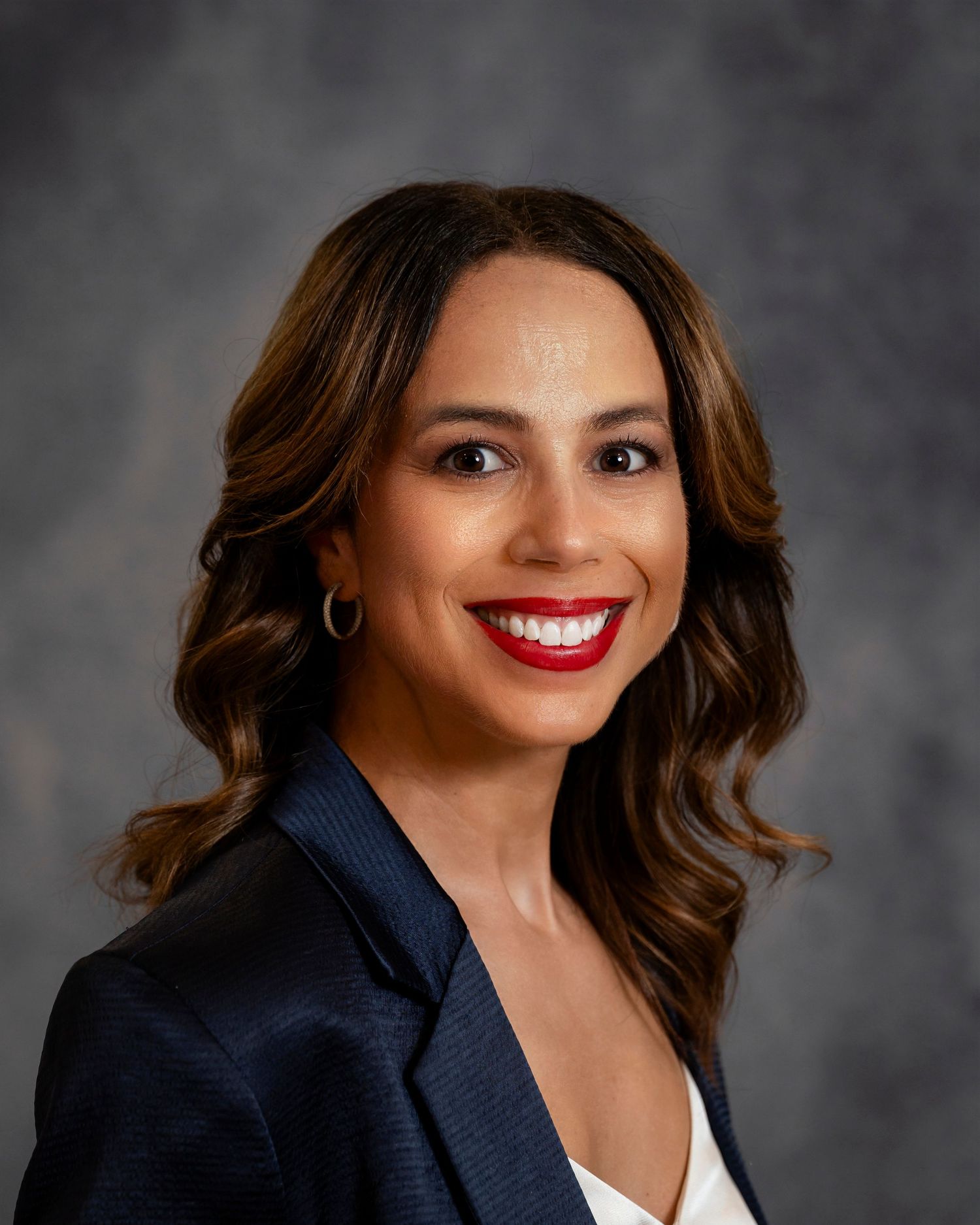 Professional headshot portrait in navy blazer with white top against gray background showing bright smile.