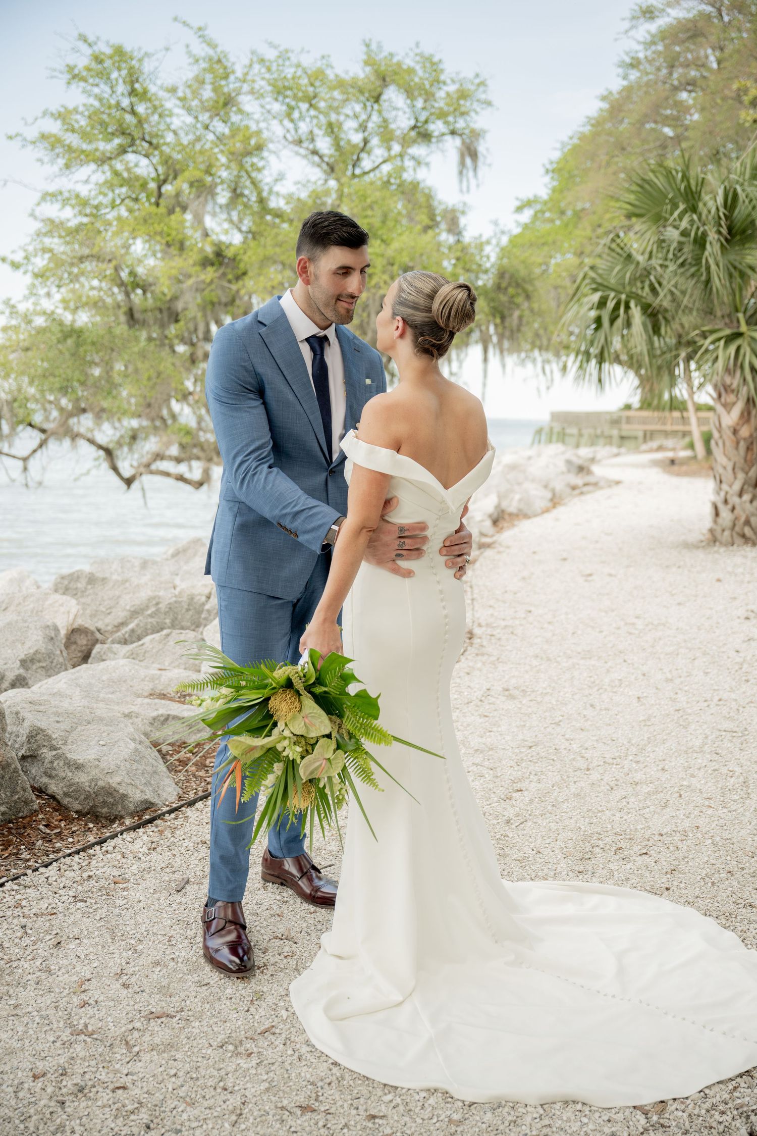 A serene beachside wedding scene with a couple in elegant attire sharing a romantic moment by palm trees and water.