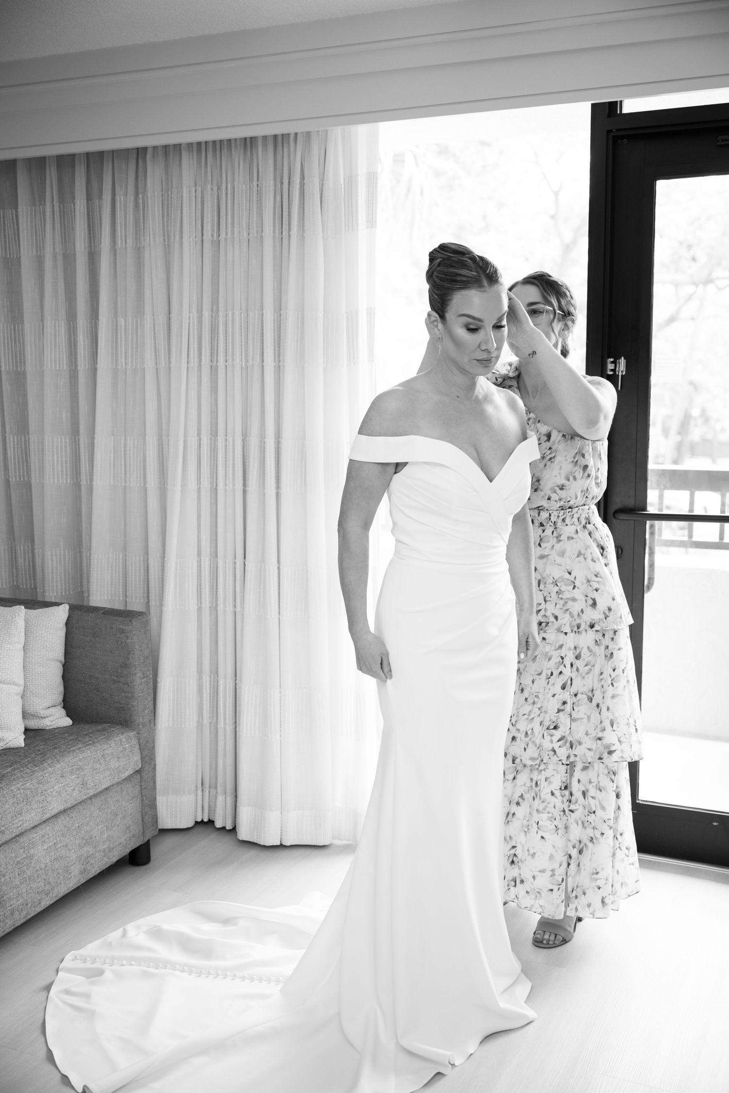 Black and white photo of a bride in an elegant off-shoulder wedding gown with train as someone assists with the dress.